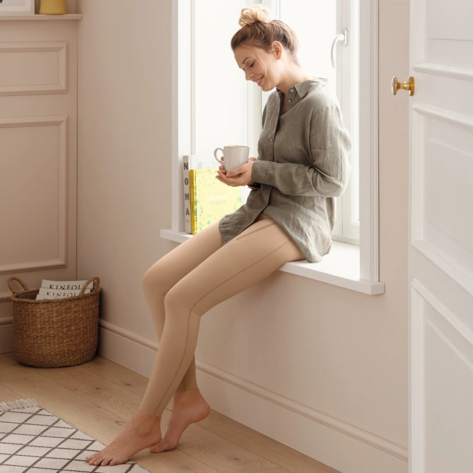 Female model sitting in front of a window at home, enjoying coffee while wearing comfortable Juzo compression pants.